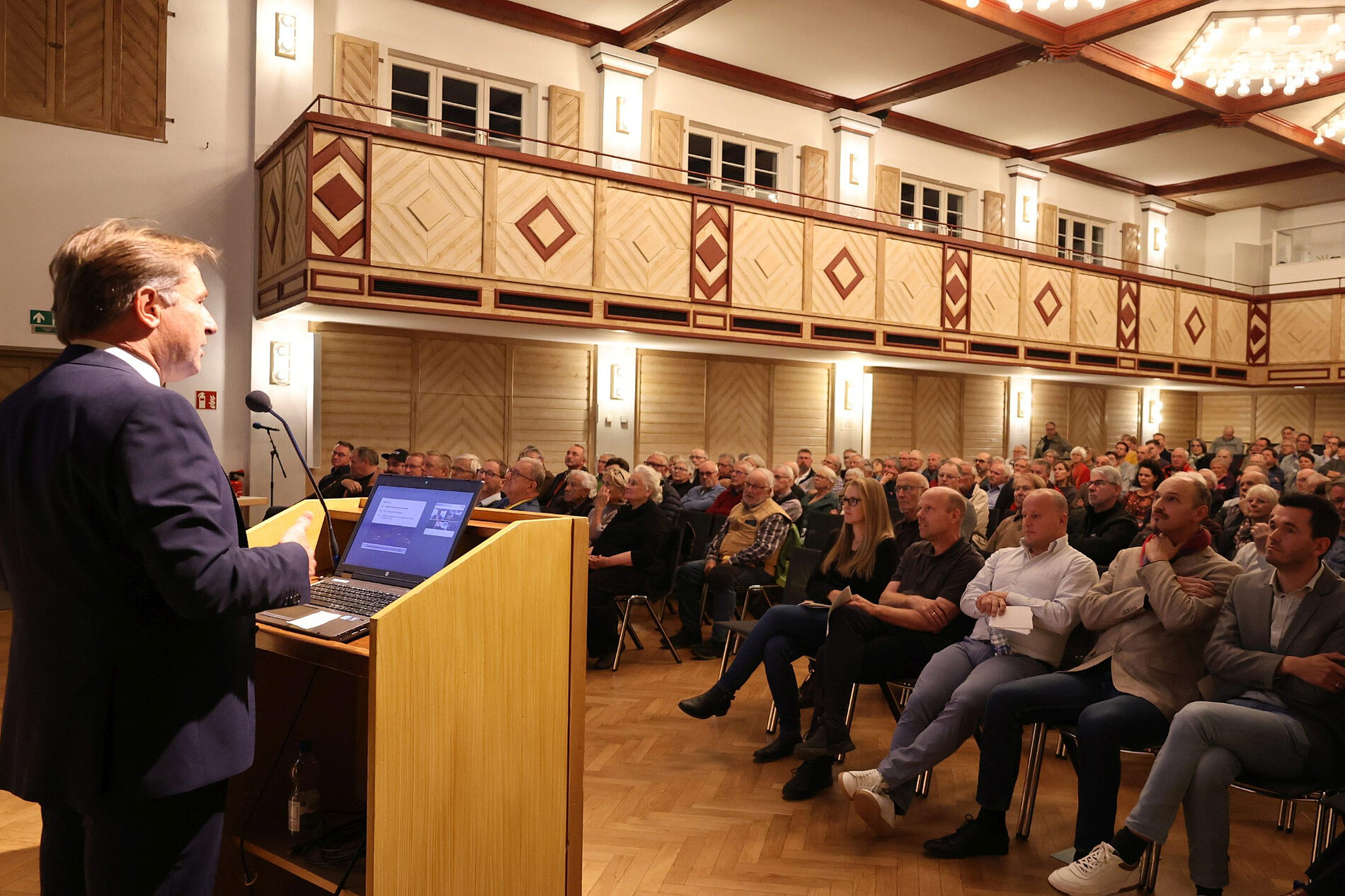 Ein Referent steht an einem Rednerpult und spricht vor einem Publikum in einem großen Saal. Die Zuschauer sitzen in Reihen auf Stühlen und sehen gebannt in Richtung des Redners. Der Raum ist elegant mit dekorativen Wänden und Beleuchtung.