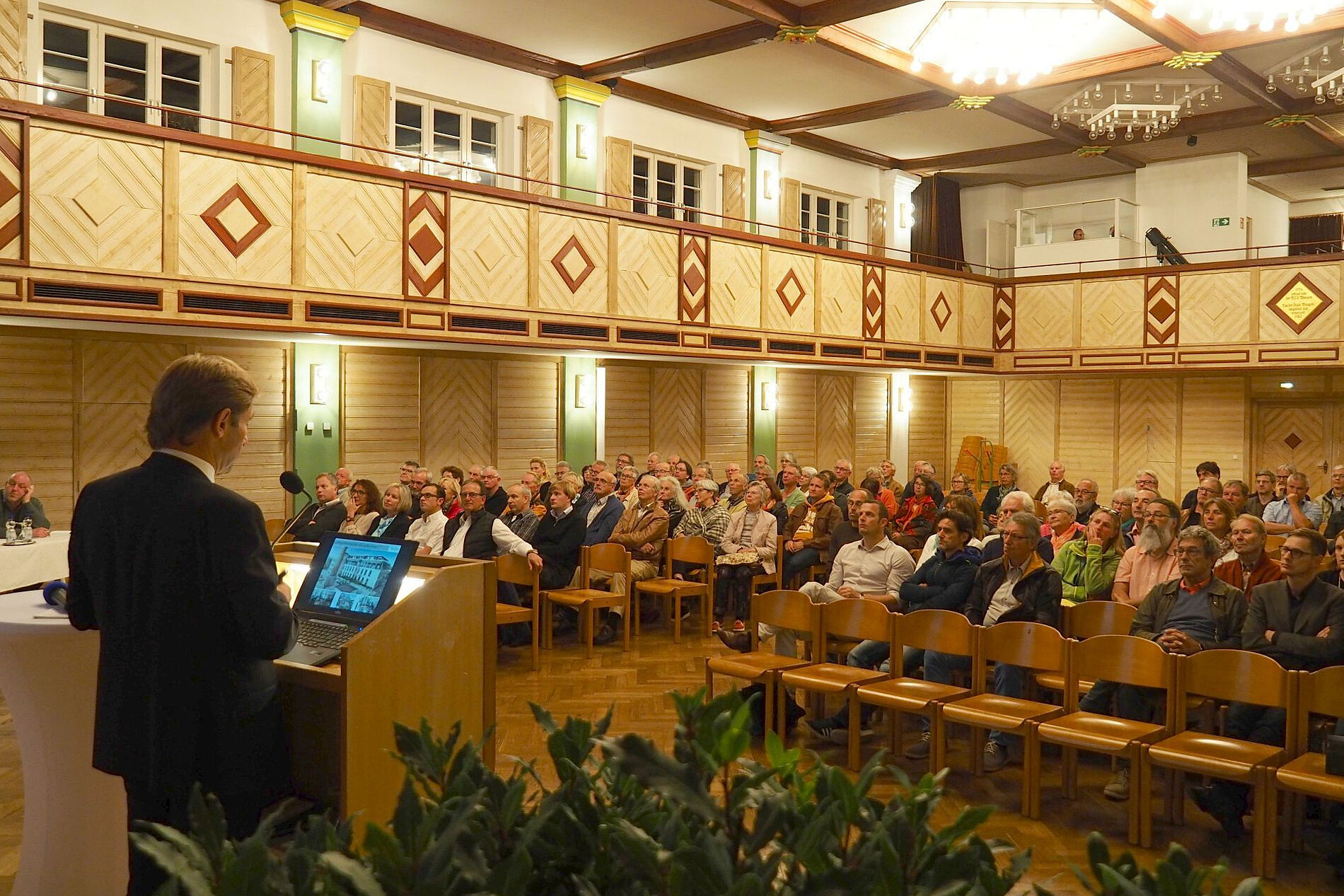 Ein Referent steht an einem Rednerpult und spricht vor einem Publikum in einem großen Saal. Die Zuschauer sitzen in Reihen auf Stühlen und sehen gebannt in Richtung des Redners. Der Raum ist elegant mit dekorativen Wänden und Beleuchtung.