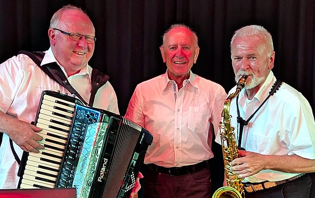 Drei Männer stehen auf einer Bühne. Der Mann links spielt ein Akkordeon, der Mann in der Mitte lächelt, und der Mann rechts hält ein Saxophon. Alle tragen helle Hemden und scheinen Musik zu machen. Der Hintergrund ist dunkel.