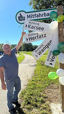 Ein Mann steht vor einem Schilderbaum, der in verschiedene Richtungen zeigt. Die Schilder weisen auf die Orte Karsee, Herfatz, Wangen und Voro hin. Der Mann zeigt mit dem Finger auf das Schild mit der Aufschrift "Mitfahrbänke". Im Hintergrund sind Bäume und eine grüne Wiese zu sehen.