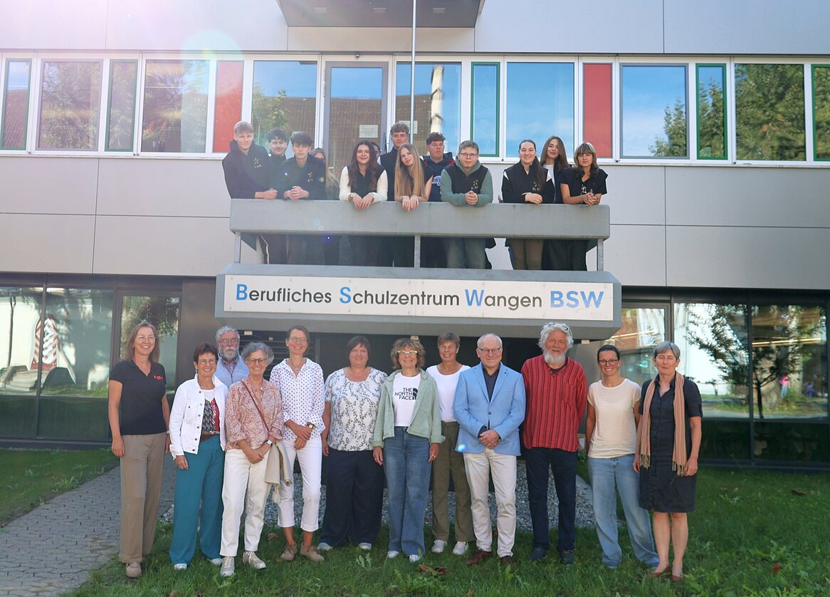Eine Gruppe von 15 Personen steht und sitzt vor einem Gebäude mit der Aufschrift "Berufliches Schulzentrum Wangen BSW". Einige Personen stehen auf einem Balkon, während andere im Vordergrund stehen. Es ist sonnig und die Atmosphäre ist freundlich.