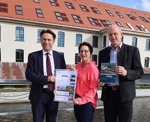 Zwei Männer und eine Frau vor der Alten Spinnerei mit dem Buch zur ERBA in der HAnd