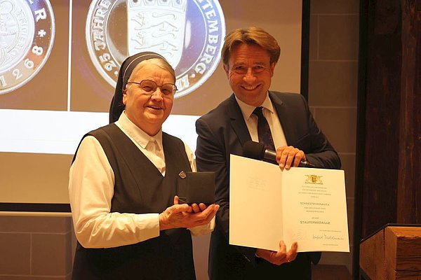 Schwester Bonavita und Oberbürgermeister Michael Lang. Beide lächeln in die Kamera. OB Lang hält die Urkunde und Schwester Bonavita die Staufermedaille in den Händen.