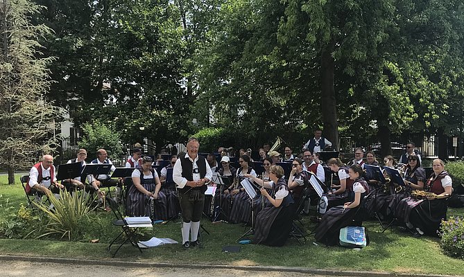 Eine Blaskapelle in traditioneller Tracht spielt im Freien auf einer Wiese. Es sind etwa dreißig Musiker zu sehen, die in Gruppen angeordnet sind. Im Hintergrund sind grüne Bäume und Sträucher. Ein Dirigent steht vorne und leitet die Musiker.