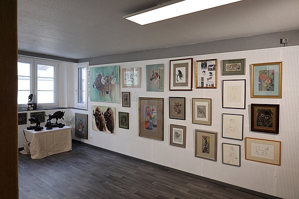 Ein Raum mit einer Wand voller gerahmter Kunstwerke, darunter Zeichnungen und Gemälde. Auf einem Tisch in der Ecke stehen einige Skulpturen. Fenster lassen natürliches Licht herein. Der Boden ist aus grauen Planken.
