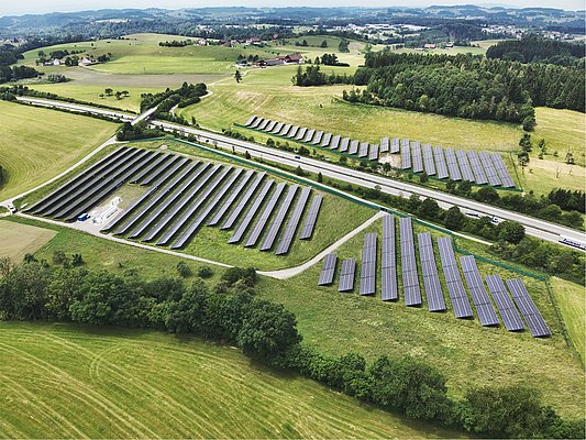 Luftaufnahme eines großen Solarparks, der in geraden Reihen angeordnete Solarpanels zeigt. Umgeben von grünen Wiesen und Bäumen, verläuft eine Straße neben dem Park. Im Hintergrund sind ländliche Gebäude und sanfte Hügel sichtbar.