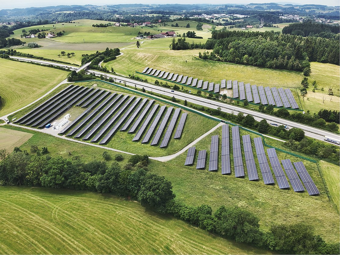 Luftaufnahme eines großen Solarparks, der in geraden Reihen angeordnete Solarpanels zeigt. Umgeben von grünen Wiesen und Bäumen, verläuft eine Straße neben dem Park. Im Hintergrund sind ländliche Gebäude und sanfte Hügel sichtbar.