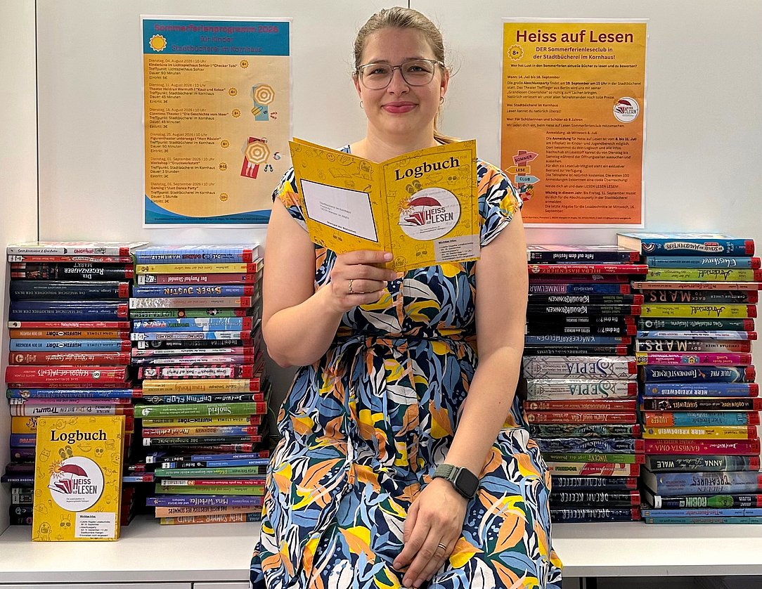 Eine Person mit Brille in einem bunten Kleid sitzt vor einem großen Bücherstapel, hält ein gelbes Logbuch in den Händen und lächelt. Im Hintergrund hängen zwei Informationsplakate darüber, wie man das Lesen fördern kann.