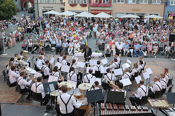 Eine große Menschenmenge sitzt auf einem Platz und schaut auf ein Blasorchester, das im Vordergrund auf einer Bühne auftritt. Die Musiker sind in weißer Kleidung und spielen verschiedene Instrumente. Im Hintergrund sind historische Gebäude und Markisen zu sehen.