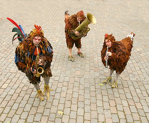 Drei Personen in aufwendigen Hühnerkostümen spielen Musikinstrumente. Die Menschen sind mit Federn und Blättern geschmückt, während sie auf einem gepflasterten Platz stehen. Ein Musiker spielt eine Klarinette, ein anderer ein Saxophon und der dritte ein Sousaphon.