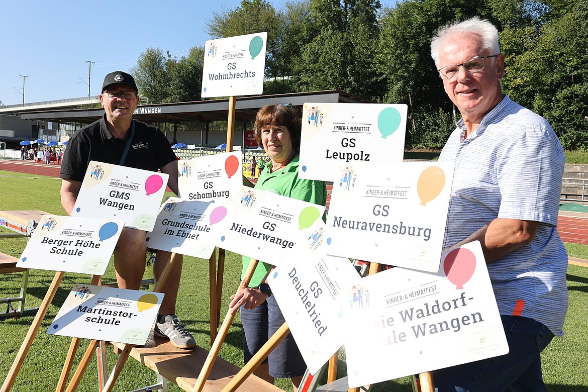 Drei Personen stehen auf einer Wiese vor Schildern mit Schulenamen, die für ein Kinderfest werben. Die Schilder beinhalten Namen wie "GMS Wangen" und "GS Neuravensburg". Im Hintergrund sind Zuschauer und eine Sporteinrichtung sichtbar.