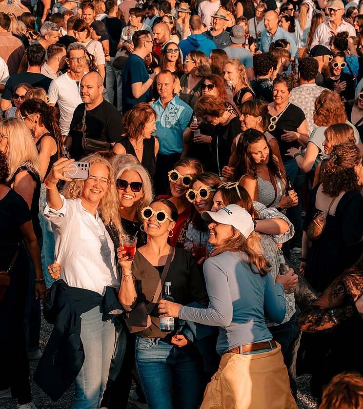 Eine Gruppe von Menschen posiert fröhlich für ein Selfie in einer belebten Menschenmenge. Einige tragen Sonnenbrillen und lächeln in die Kamera. Im Hintergrund sind viele weitere Personen zu sehen, die ebenfalls an der Veranstaltung teilnehmen.