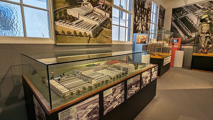 Eine Modellansicht eines historischen Geländes in einem Museum. Die Vitrine zeigt Detaildarstellungen von Gebäuden und Anlagen, umgeben von Informationstafeln und historischen Fotos an den Wänden. Helle Fenster sorgen für ausreichend Licht im Raum.