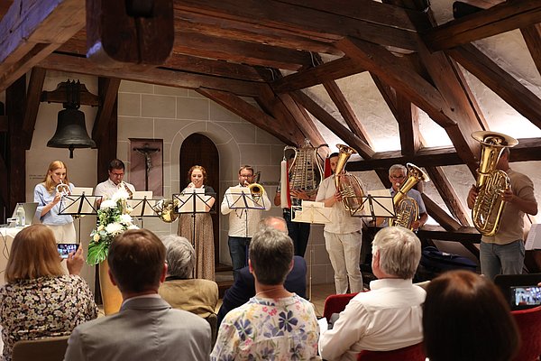Eine Gruppe von Musikern spielt auf der Bühne in einem Raum mit Holzbalken. Sie halten ihre Notenständer und spielen verschiedene Blasinstrumente, während das Publikum sitzend zuhört. Im Hintergrund ist ein großer Glockenstuhl sichtbar.