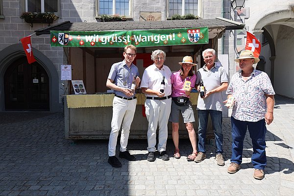 Sechs Personen stehen vor einem Verkaufsstand mit dem Schild „Wir grüßt Wangen!“ im Freien. Sie halten kleine Flaschen oder Objekte in den Händen und tragen Sommerbekleidung. Im Hintergrund sind eine historische Gebäudefront und Schweizer Flaggen sichtbar. Es scheint ein sonniger Tag zu sein.