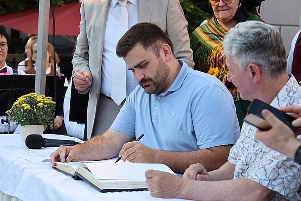 Ein Mann in einem blauen Poloshirt sitzt an einem Tisch und schreibt in ein Buch. Neben ihm sitzt ein älterer Mann. Im Hintergrund sind weitere Personen und Pflanzen zu sehen. Es scheint sich um eine feierliche Veranstaltung zu handeln.