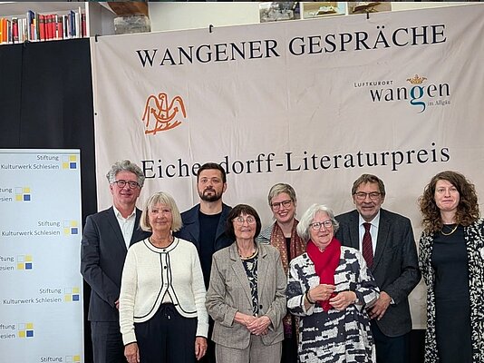Eine Gruppe von sieben Personen steht vor einem Banner mit der Aufschrift "Wangener Gespräche" und "Eichendorff-Literaturpreis". Die Personen lächeln und sind in formeller Kleidung gekleidet. Im Hintergrund sind Regale mit Büchern zu sehen.