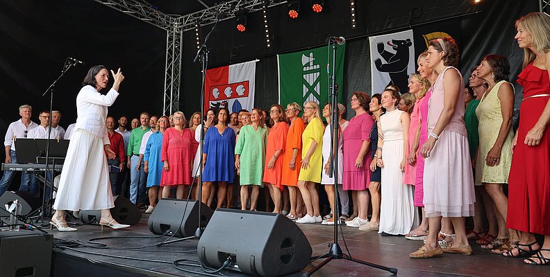 Eine Gruppe bunt gekleideter Frauen und Männer in weißen Hemden und Jeans singen.
