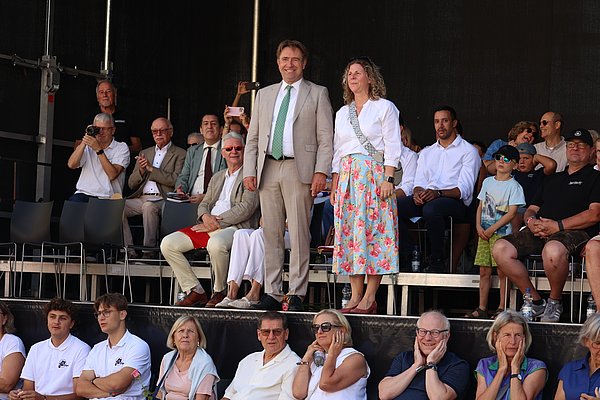 Eine Frau in einem Blumenrock und ein Mann in Anzug stehen zusammen auf einer Bühne. Im Hintergrund sitzen zahlreiche Zuschauer, einige applaudieren oder schauen aufmerksam. Die Stimmung ist feierlich und interessiert.