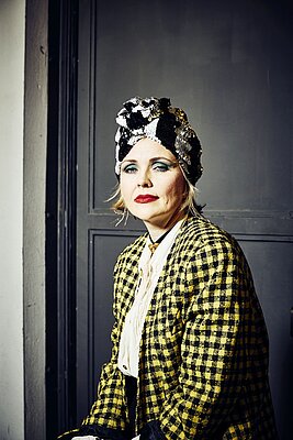 Eine Person mit einem auffälligen karierten Outfit und einem glitzernden Kopfschmuck sitzt vor einer grauen Wand. Sie hat auffällige Make-up-Farben, darunter rote Lippen und starke Augenbetonung.