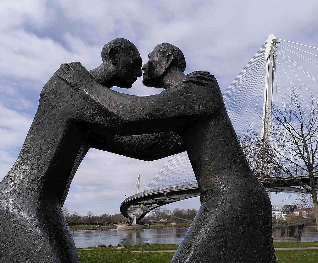 STatue mit zwei Menschen, die sich umarmen. Im Hintergrund sieht man die Europabrücke in Kehl