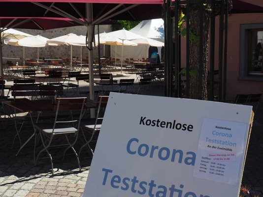 Nahaufnahme eines Informationsschilds zur Teststation an der Eselsmühle sowie Außengastronomiebestuhlung und der Teststation im Hintergrund