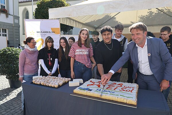 Eine Gruppe von Menschen steht an einem Tisch mit einem großen Kuchen und Cupcakes. Ein Mann in Anzug schneidet den Kuchen, während mehrere Jugendliche und eine Frau daneben stehen. Im Hintergrund ist ein Zelt und ein Banner mit dem Text „Weltladen El Sol“.