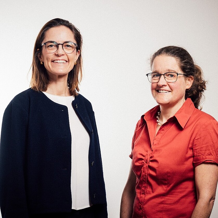 Zwei Frauen stehen nebeneinander vor einem hellen Hintergrund. Die links trägt eine dunkle Jacke und eine helle Bluse, die rechts eine rote Bluse und wirkt freundlich. Beide Frauen tragen Brillen und lächeln in die Kamera.