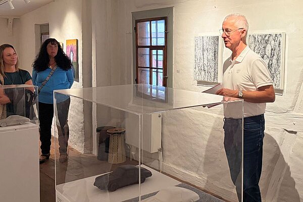 Eine Person präsentiert in einem Ausstellungsraum Kunstwerke. Zwei weitere Personen hören aufmerksam zu. Der Raum hat helle Wände und ist mit Kunstwerken dekoriert. Eine transparente Vitrine steht im Vordergrund, in der sich ein graues Objekt befindet.